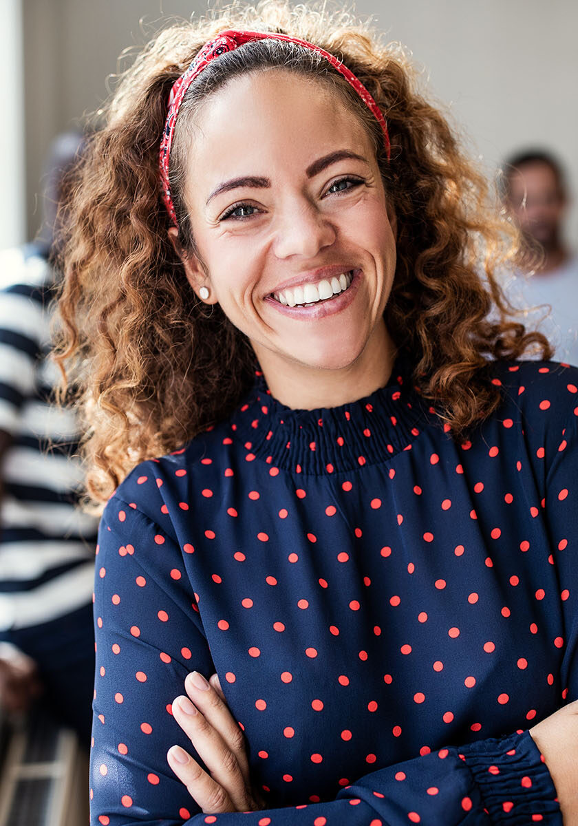 Young female designer smiling while standing with her arms crossed after an office meeting with colleagues standing in the background